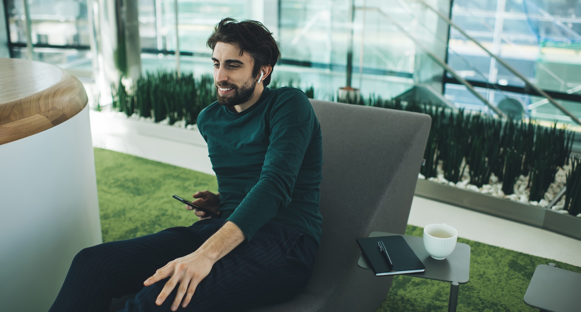 Cheerful young man having phone conversation via TWS earbuds sitting on lounger